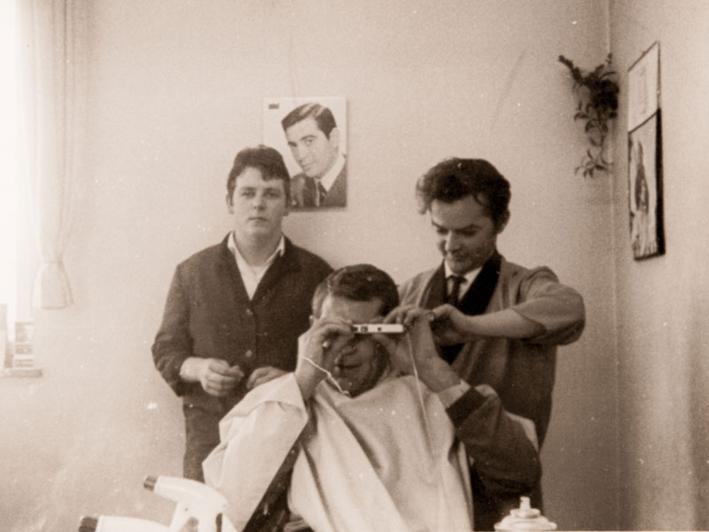 Walter Penkalla in den 60er beim Haareschneiden eines Kunden, der Kunde macht das Foto über den Spiegel. Im Hintergrund Stefan Penkalla